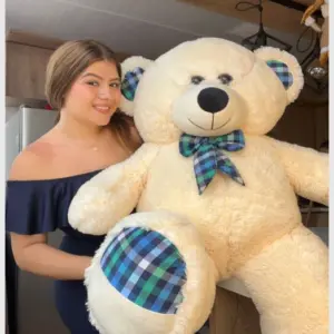 Oso de peluche gigante Caramelo de 145 cm en color beige, con lazo y parches en cuadros azules y verdes, abrazado por una mujer.